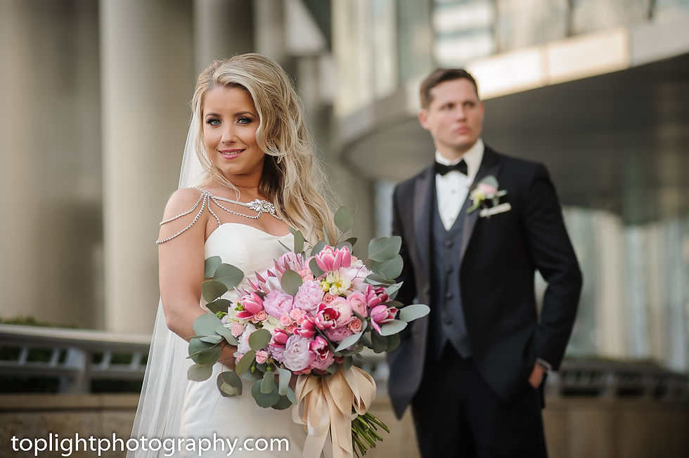 Bride in white dress holding pink bouquet, smiling. Groom in black tuxedo in background. Urban setting. Websites text: toplightphotography.com.