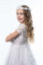 Young girl in white first communion lace dress and floral headpiece, smiling with arms crossed. Background is a plain light color, creating a joyful mood.