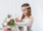 Girl in white first communion dress reads a book, wearing a floral headband. Pink and white flowers beside her. Bright, soft-lit background.