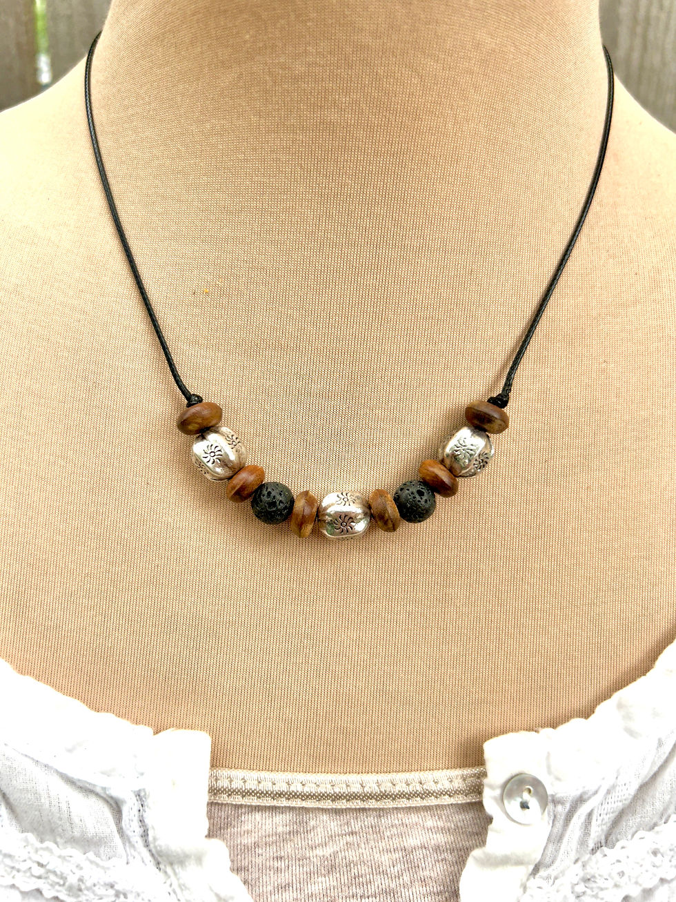 Black 1mm faux leather cord necklace with 3 stamped silver beads, 6 wood rondelles, and 2 porous black lava beads displayed o