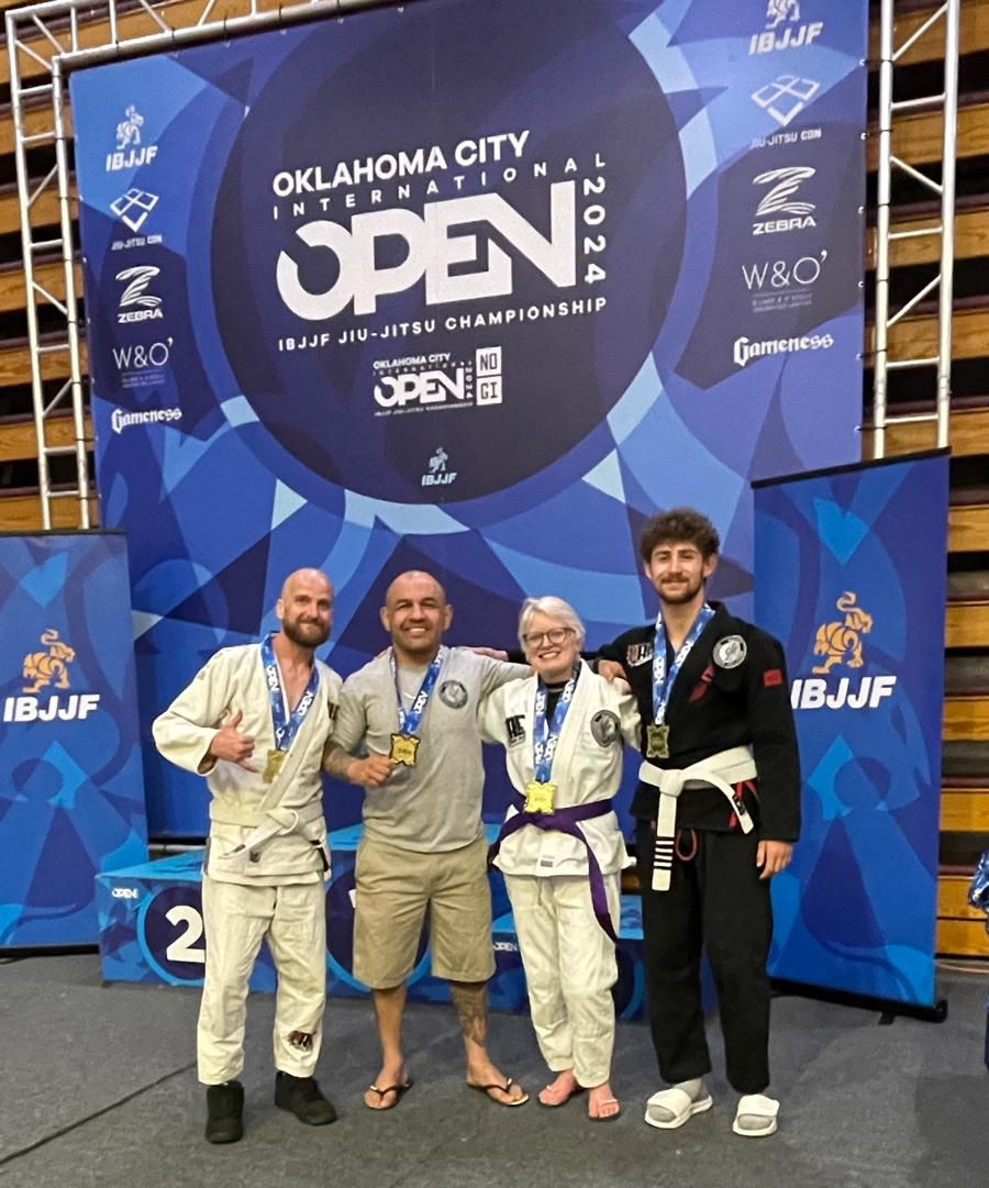 Four people with medals pose happily in front of an "Oklahoma City Open 2024" Jiu-Jitsu banner. They're wearing martial arts attire.
