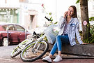 Smiling young woman sitting on a stone bench outdoors, talking on the phone next to a white bicycle with a basket, on a sunny day in the city.
