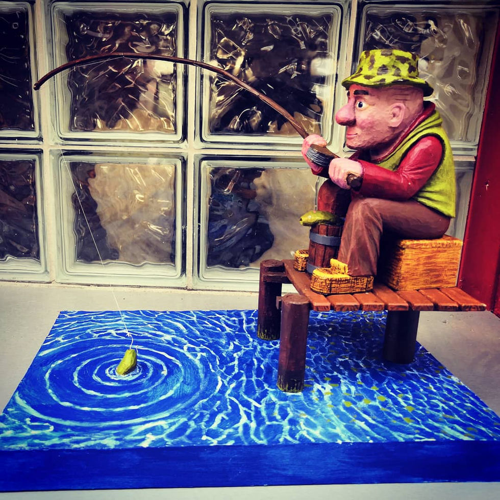 ca 30 cm große, geschnitzte und bemalte Figur eines Anglers auf einem Steg. Auf einer Kiste sitzend, mit der Angel in der Hand und einem Eimer zwischen den Beinen, in dem bereits ein gefangener Fisch liegt