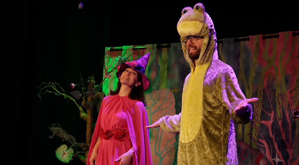 Zdjęcie ze spektaklu teatralnego dla dzieci “Wesoła Czarownica”. Na pierwszym planie aktor w stroju Żaby. Ma rozłożone ręce. Uśmiecha się. Na drugim planie aktorka w różowym szpiczastym kapeluszu i różowym stroju. Uśmiecha się. W tle kolorowe drzewa. 