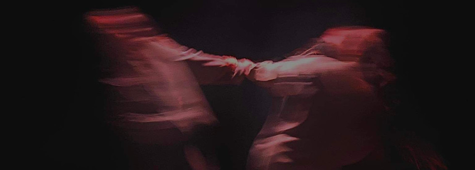 Dwie niewyraźne, rozmyte sylwetki trzymające się za ręce, na oświetlonej scenie. Wszystko w fioletowo z bordowych kolorach. Zdjęcie przykryte napisem “Square. Warsztaty taneczne dla młodzieży i dorosłych”. “Square” - napisane jest większa, żółta czcionką. Dalsza część napisu, ma mniejszą czcionkę i czarne litery. Zapraszamy na taniec współczesny w Warszawie.