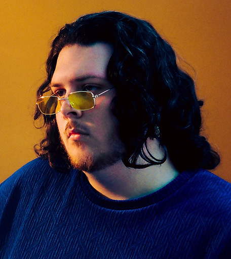 Portret długowłosego młodego mężczyzny w żółtych okularach i niebieskiej koszulce na pomarańczowym tle.