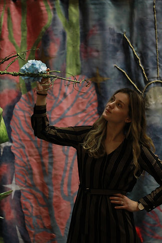 Młoda kobieta w czarno-brązowej sukience w paski trzyma w ręku gałąż zielonego drzewa ze scengrafii spektaklu dla dzieci Wesoła Czarownica.