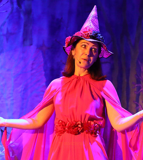 Zdjęcie ze spektaklu teatralnego dla dzieci “Wesoła Czarownica”. Około 50letnia kobieta, w różowym stroju i różowym, szpiczastym kapeluszu, stoi z rozłożonymi rękami, w fioletowym świetle. Występujemy z naszym edukacyjnym teatrem dla szkół.