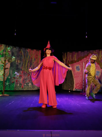 Zdjęcie ze spektaklu teatralnego dla dzieci “Wesoła Czarownica”. Na pierwszym planie aktorka w szpiczastym różowym kapeluszu I różowej sukience. Śpiewa i tańczy. W tle tańczy aktor w stroju Żaby. Z tyłu namalowany na płótnie różowo - zielono - niebieski las i domek. Po lewej stronie zielone drzewo. Dynamiczna scena z naszego przedstawienia dla najmłodszych.