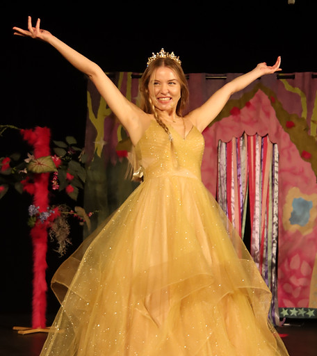 Zdjęcie ze spektaklu teatralnego dla dzieci “Wesoła Czarownica”. Młoda kobieta w złotej sukience, uśmiecha się i rozkłada do góry ręce. W tle namalowany na płótnie różowo - niebieski domek i różowo - zielone drzewo. Scenografia naszego teatru objazdowego dla dzieci.