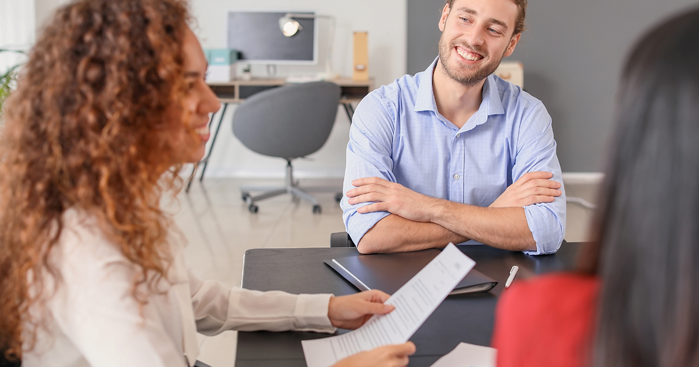A group of coworkers sit at a table in a meeting. They are all smiling. A woman leading the group holds a piece of paper important to the meeting in her hands. Around them is a corporate office setting.