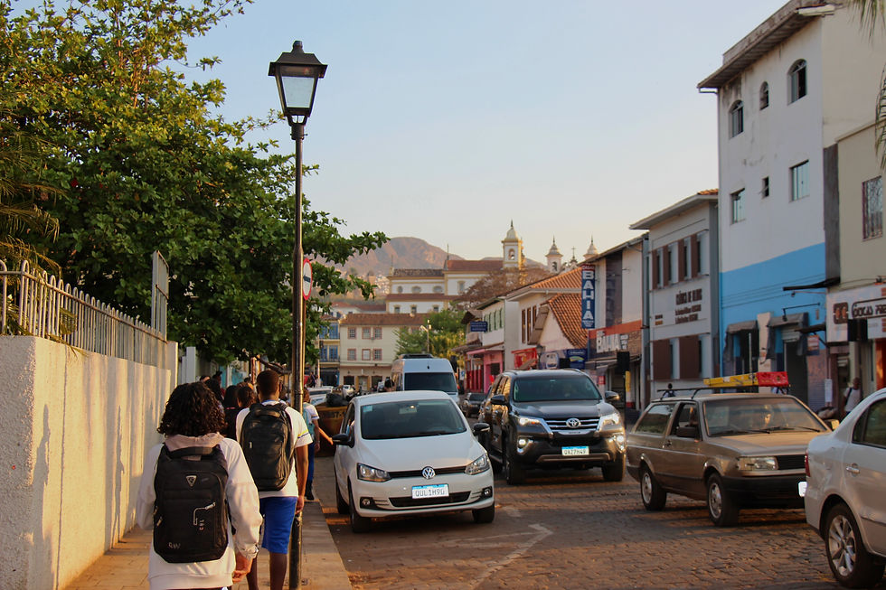 #paratodosverem: em primeiro plano, lado esquerdo da imagem, dois estudantes com suas mochilas pretas caminham ao lado de um muro branco, no centro da cidade, todos cobertos pela luz do sol em uma calçada aparentemente cheia de outros pedestres do centro ao do lado direito da foto há uma extensa fila de carros, nas cores branca, preta e cinza. Ao fundo, sobrados, igrejas e lojas. Na porção superior é possível ver um céu azul bem esbranquiçado.
