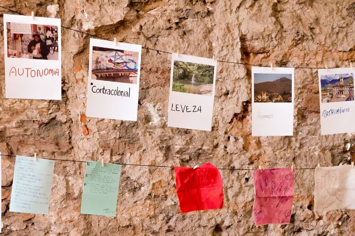 #paratodosverem: A imagem representa três varais com os trabalhos dos participantes; no primeiro observa-se imagens de cidades históricas por onde o programa passou. O segundo e o terceiro há papeis e pedaços de tecidos de cores diferentes e com escritos e desenhos feitos pelos participantes. Ao fundo é possível ver uma parede de pedras.
