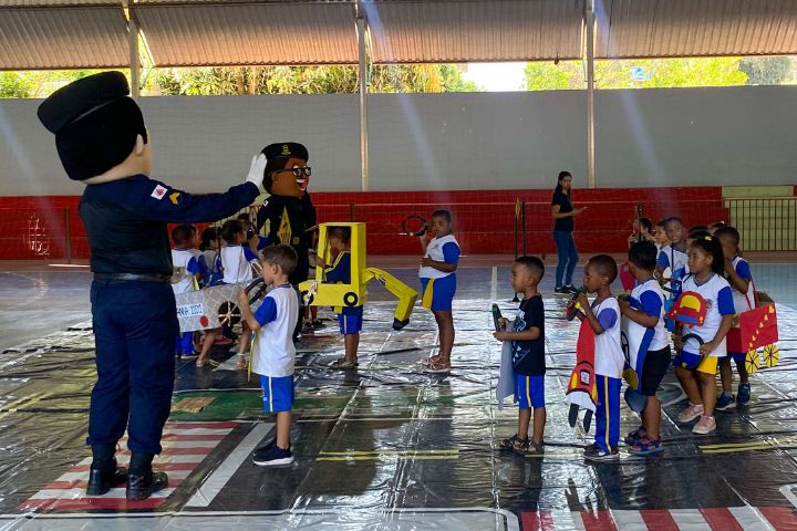 #Paratodosverem: Na imagem os alunos estão em uma quadra, nós vemos um tapete que simula o trânsito de uma cidade, contendo avenidas e faixas de pedestres. Sobre ele, há algumas crianças com uniforme escolar, sendo ele branco e azul, e estão segurando pequenos volantes, com imitações de carros feitos de papelão. As crianças estão enfileiradas uma atrás da outra, seguindo o mascote masculino do Demutran, enquanto a mascote feminina fecha a fila.