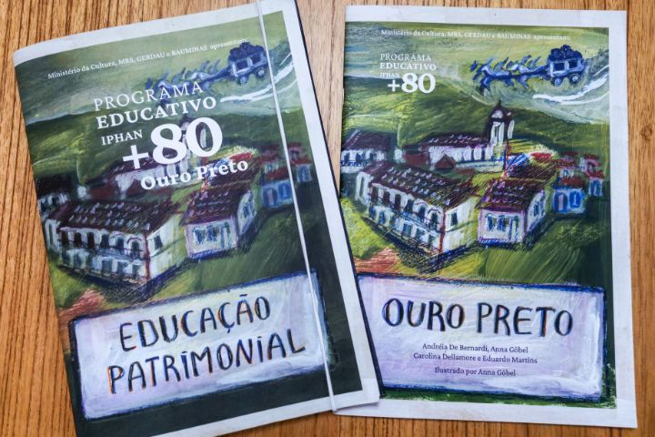 #paratodosverem: A imagem mostra dois cadernos sobre uma mesa de madeira. À esquerda, está o caderno com o escrito em letras grande e em destaque na parte superior: “Programa Educativo Iphan +80”, seguido por “Educação Patrimonial”. Ao fundo, há uma pintura que retrata a cidade de Ouro Preto, e na parte superior, uma ilustração de uma carruagem. À direita, encontra-se o outro caderno, que apresenta o escrito em letras pequenas e em destaque na parte superior “Programa Educativo Iphan +80” e, logo abaixo, “Ouro Preto”. Ao fundo, é possível ver a mesma imagem do caderno anterior.
