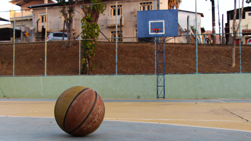#ParaTodosVerem - Em primeiro plano, uma bola de basquete laranja mais à esquerda sobre o piso azul e amarelo com algumas rachaduras da quadra. Em segundo plano, uma tabela de basquete azul escura com o aro laranja sem rede. Casas, carros e árvores desfocados mais ao fundo.