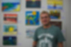 #ParaTodosVerem: Homem de pele clara, cabelos loiros cacheados e curtos, usa óculos e uma camiseta verde com a estampa. Ele está sorrindo e posando em frente a uma parede com várias pinturas identificadas com etiquetas. As obras mostram diferentes paisagens como praias, desertos e montanhas.