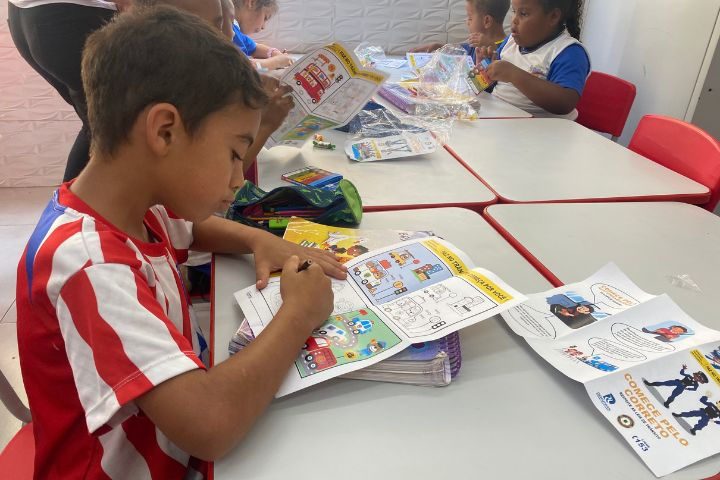 #Paratodosverem: Na imagem, vemos seis crianças sentadas. Algumas estão com uniforme da escola, e uma delas está com uniforme de futebol. Sobre as mesas, há estojos e apostilas educativas para colorir. Algumas crianças estão pintando essas apostilas. As mesas são brancas com bordas vermelhas.