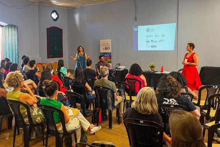 #paratodosverem: A imagem destaca em primeiro plano os participantes do Programa, que estão sentados de costas para a câmera. É possível observar um público diversificado, predominantemente formado por mulheres. Ao fundo, estão os idealizadores do Programa: à esquerda, em pé, Andréia De Bernardi; ao seu lado, sentado, Eduardo Martins; e à direita da fotografia, em pé, Carolina Dellamore.