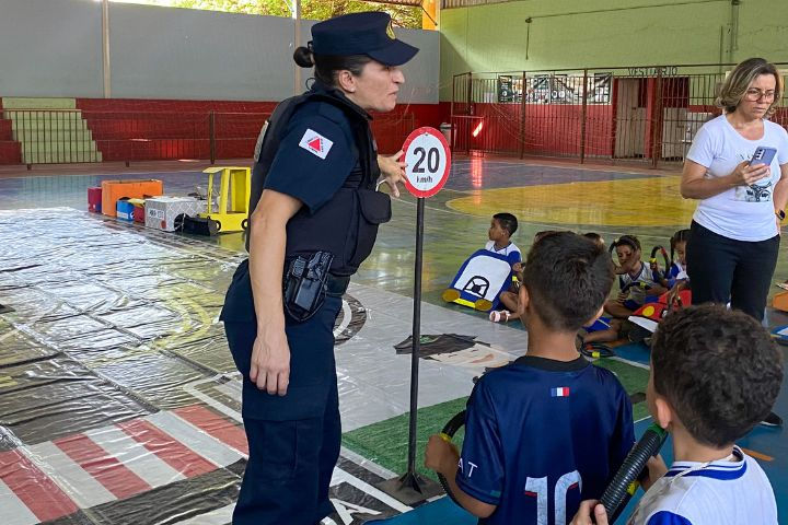 #Paratodosverem: Na imagem, vemos algumas crianças segurando volantes de simulação, no canto inferior direito . Uma das crianças está de uniforme e outra com uma camisa de futebol. Na frente delas, há uma guarda do Demutran apontando para uma placa. No lado esquerdo, há mais um grupo de estudantes sentados na quadra, acompanhados por uma professora, e ao lado deles, há carros feitos de papelão.
