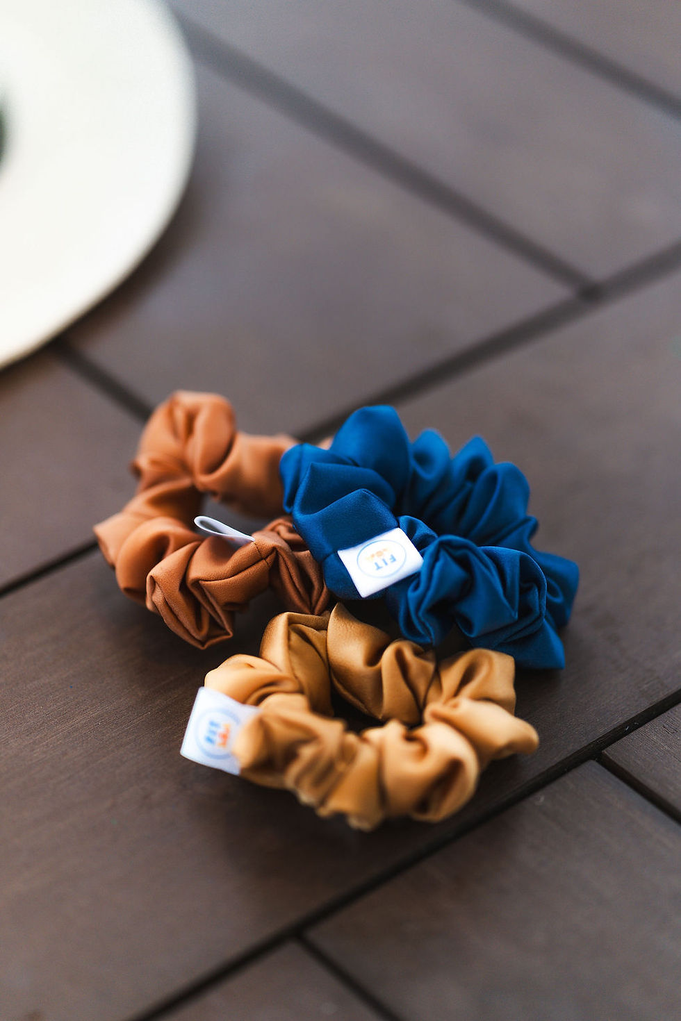 Close up of FIT30A scrunchies in Coastal Blue, Golden Hour Orange, and Sunkissed Yellow on a wooden table in 30A Florida, per