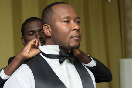 Groom in tuxedo adjusting his bow tie with assistance.