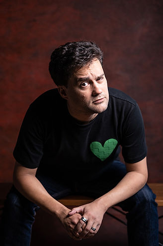 You're looking at Tony Johnson, a thoughtful looking man with curly brown hair, a rather cheeky pensive expression, wearing a black T-shirt with a green love heart on it. He's leaning towards the viewer looking quisical. This is a frequent Tony Johnson expression because he is a scientist who questions everything all the time.