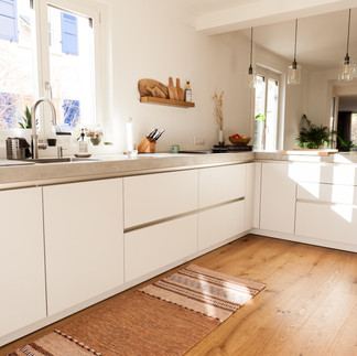 Bright, minimalist kitchen with white handleless cabinets, concrete countertops, light wood flooring, and sunlight streaming through a window.