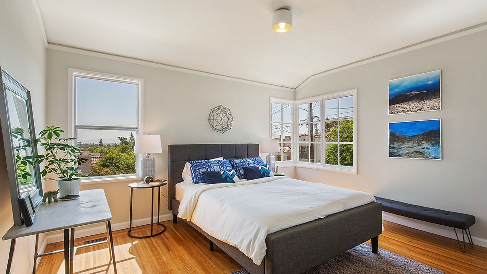Bright bedroom with a large bed, blue pillows, and wood floor. Windows show a sunny view. Art and plants add a cozy touch.