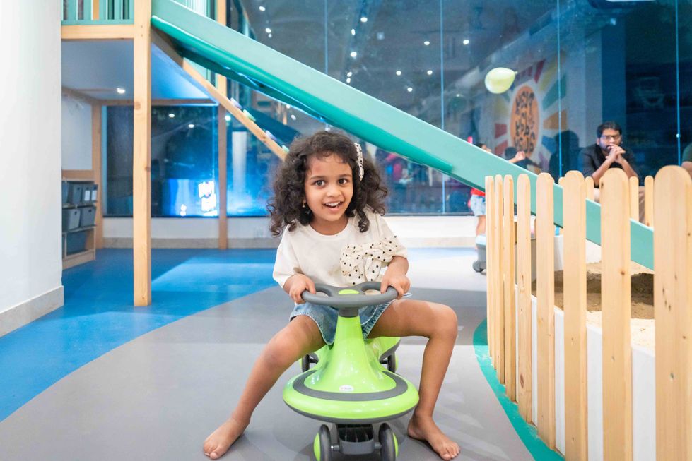 Child playing on a toy car indoors, smiling.