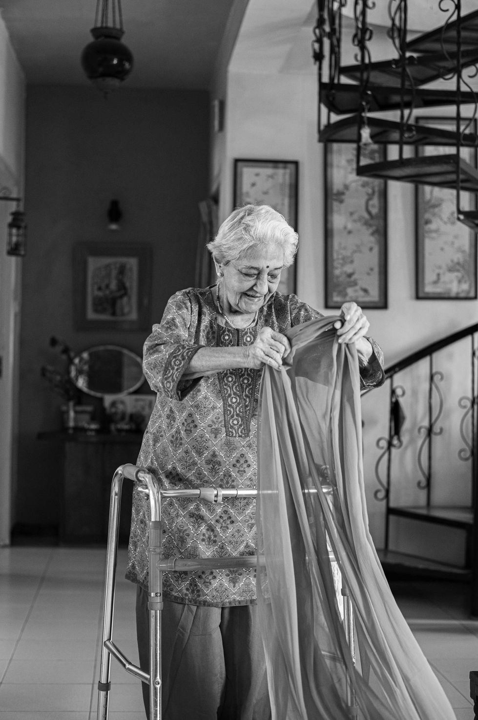 Elderly woman smiling, holding fabric, near staircase.