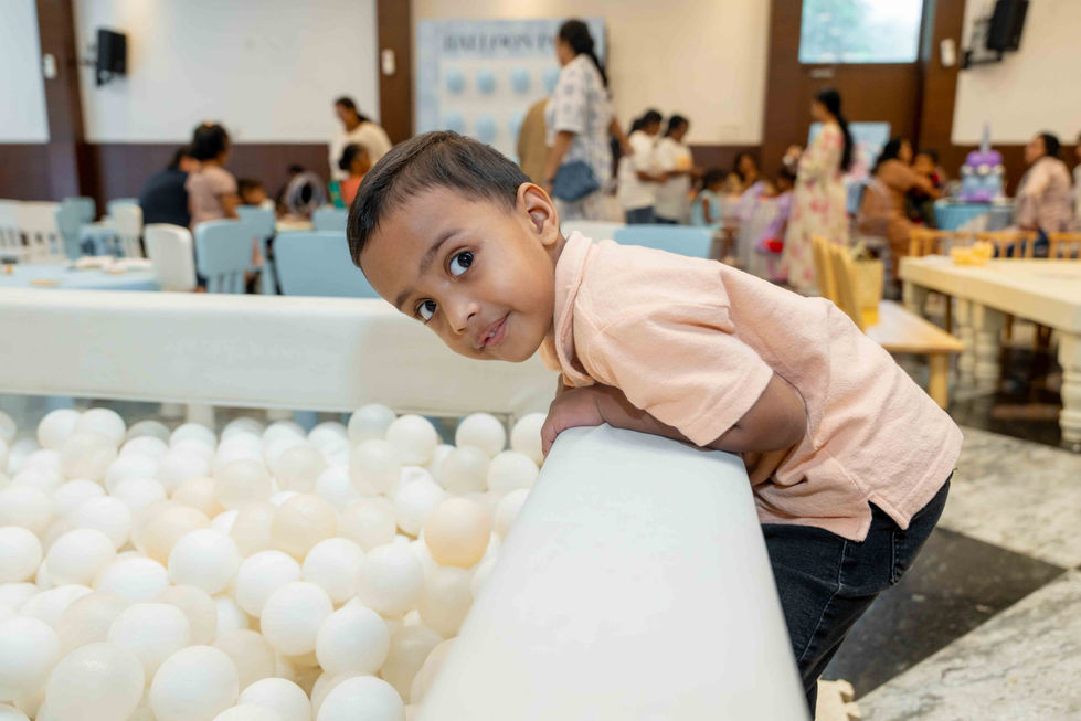 Child leaning over ball pit, people in background.