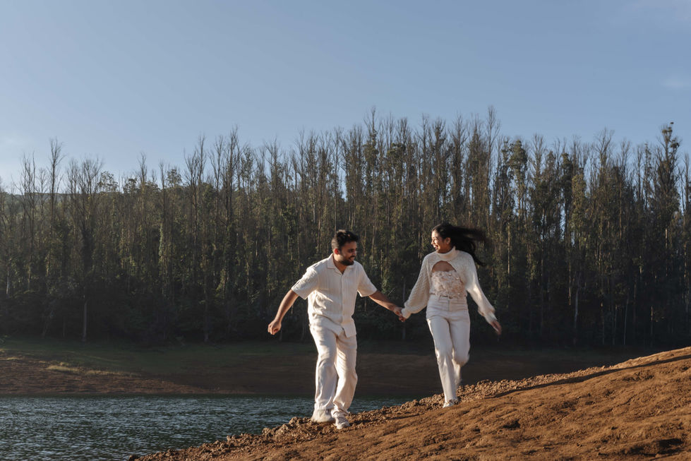 Couple in white walking near a lake, forest background.