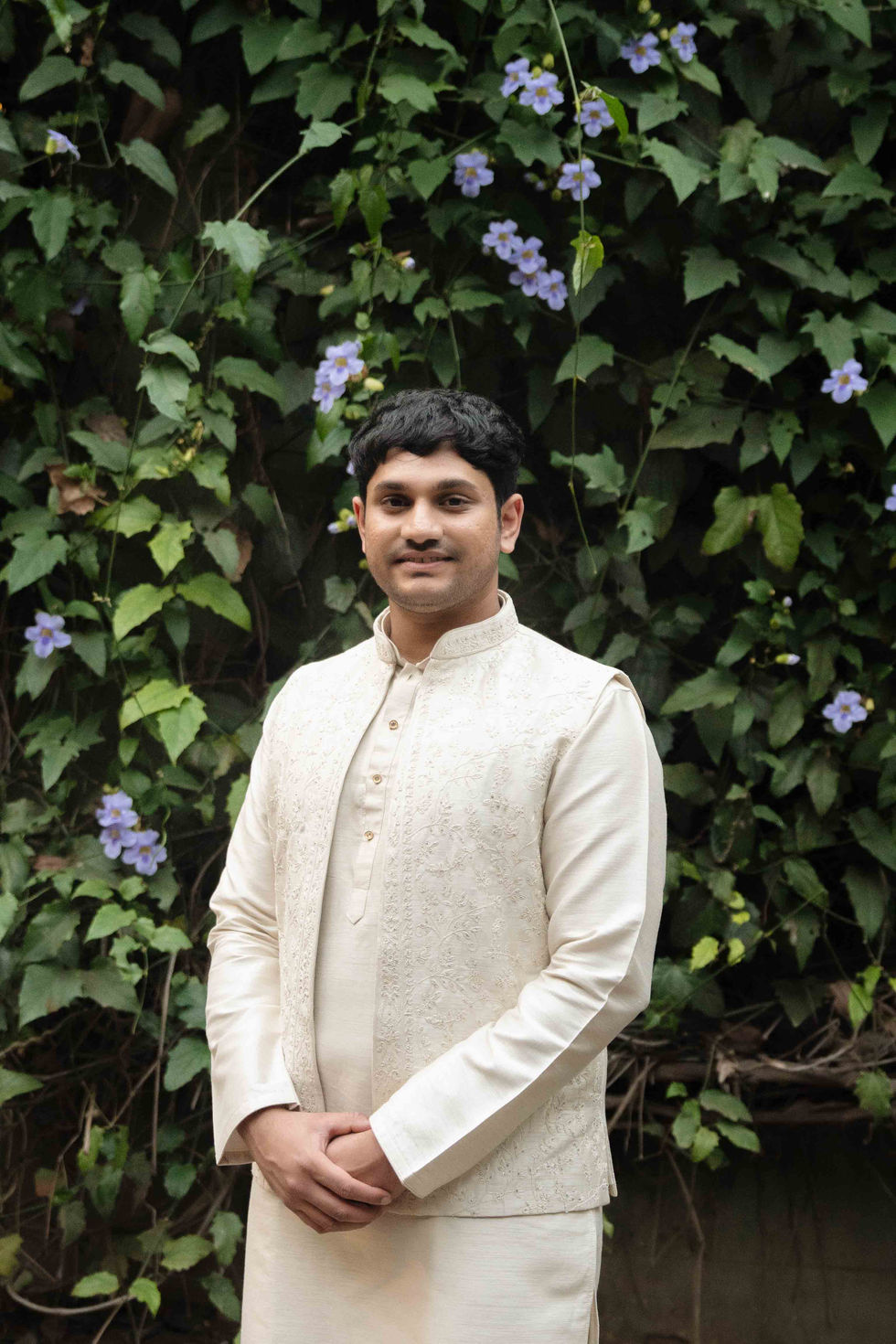 A person in traditional attire, standing before foliage.