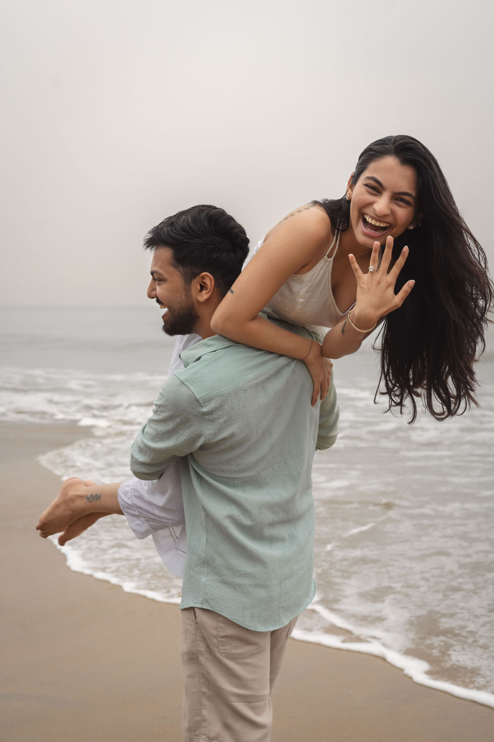 Man carrying his fiancé at the beach while she shows her engagement ring to camera.
