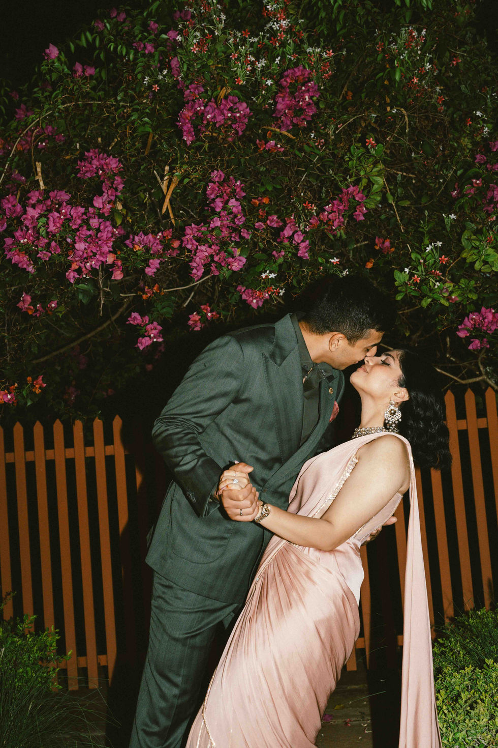 Couple kissing in garden, surrounded by pink flowers.