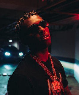 Foto de Caio Luccas, um homem usando óculos escuros, blusa preta e grandes cordões de ouro, iluminado por uma luz vermelha.