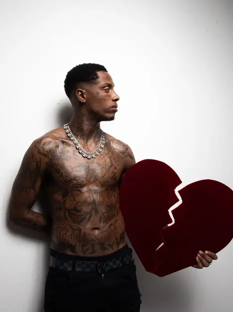 Foto de Kayblack, um homem tatuado e sem camisa, segurando um coração partido vermelho.