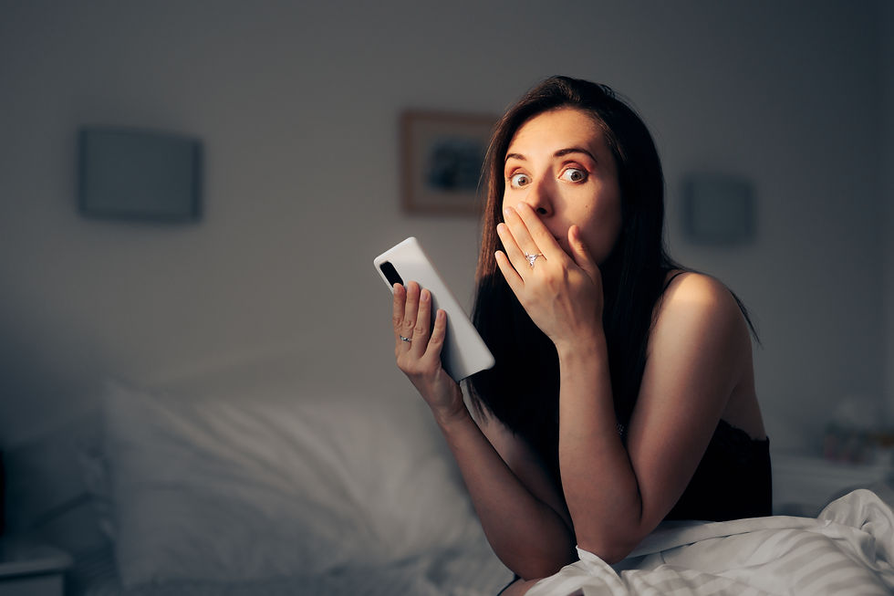 A surprised woman in a dimly lit room covers her mouth in disbelief while reading an intimate message on her phone.
