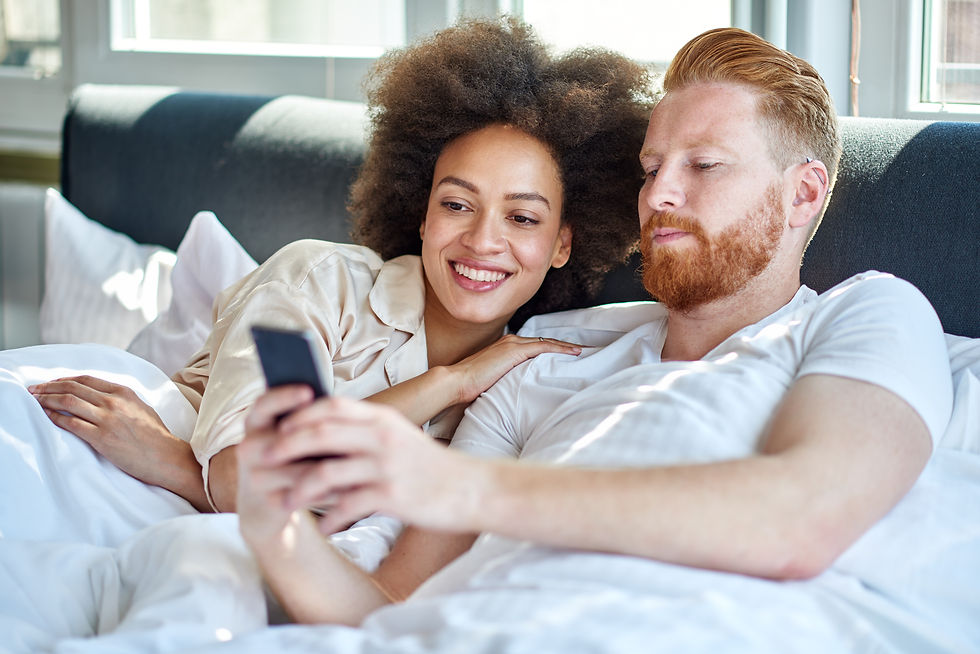Couple lounging in bed, smiling and looking at a smartphone. Brightly lit room with white bedding, creating a cozy and relaxed atmosphere.
