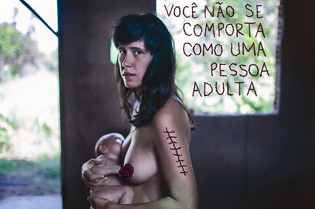 Fotografia artística de uma mulher de pele clara e cabelos escuros, segurando um bebê nos braços dentro de um espaço inacabado ou abandonado, com uma janela aberta ao fundo que revela a vegetação externa. Ela está sem roupa na parte superior do corpo e tem um seio coberto por uma aplicação em crochê vermelho, enquanto o outro é exposto para amamentação. No braço, há uma cicatriz bordada em linha vermelha, remetendo a uma sutura. Seu olhar é sério e direcionado para a câmera. Na parede ao fundo, está escrito em letras bordadas: 'VOCÊ NÃO SE COMPORTA COMO UMA PESSOA ADULTA'. A composição e os elementos bordados no corpo e no ambiente sugerem uma reflexão sobre maternidade, feminilidade e imposições sociais. Obra de Malu Teodoro, intitulada 'Você está morta'.