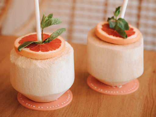 Zwei frische junge Kokosnüsse nebeneinander mit einer frischen Scheibe Grapefruit und frischer Minze als Deko mit weißem Strohalm. beide liegen auf einem pinken Untersetzer mit weißer Aufschrift.
