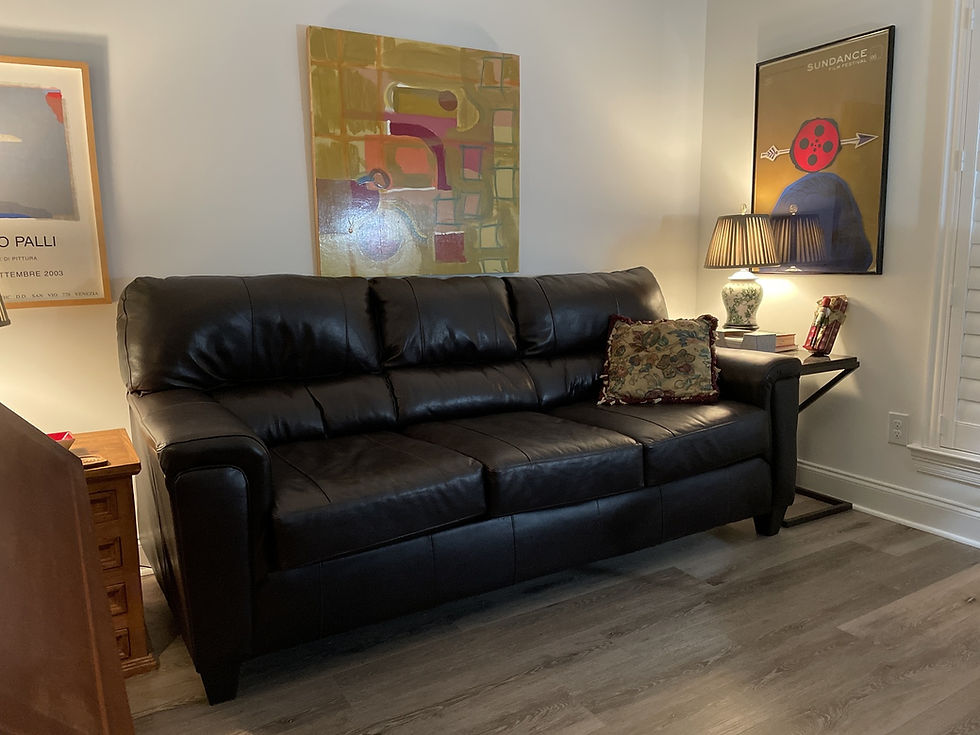 Wide landscape photo of a cozy therapy office with a black leather couch, warm lighting, and abstract art on the wall, suitable as a welcoming hero background.