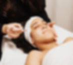 A woman lying on a spa bed with her hair wrapped as a face cream is spread across her cheeks.
