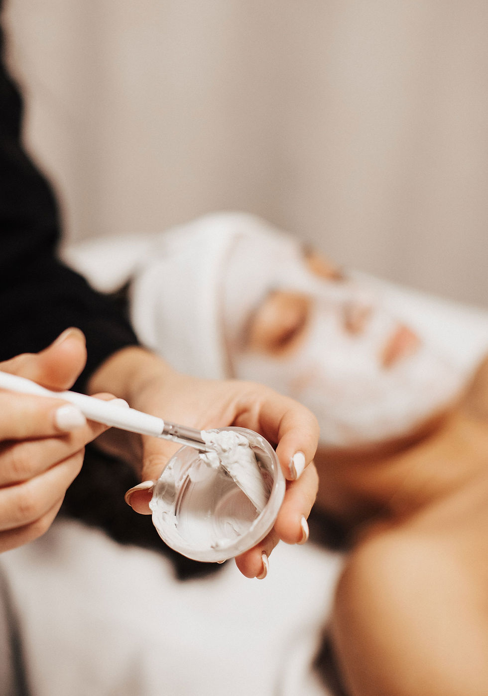 A hand holding a small bowl containing a white paste while she wipes a brush in the cream, with a woman getting a facial in the background.