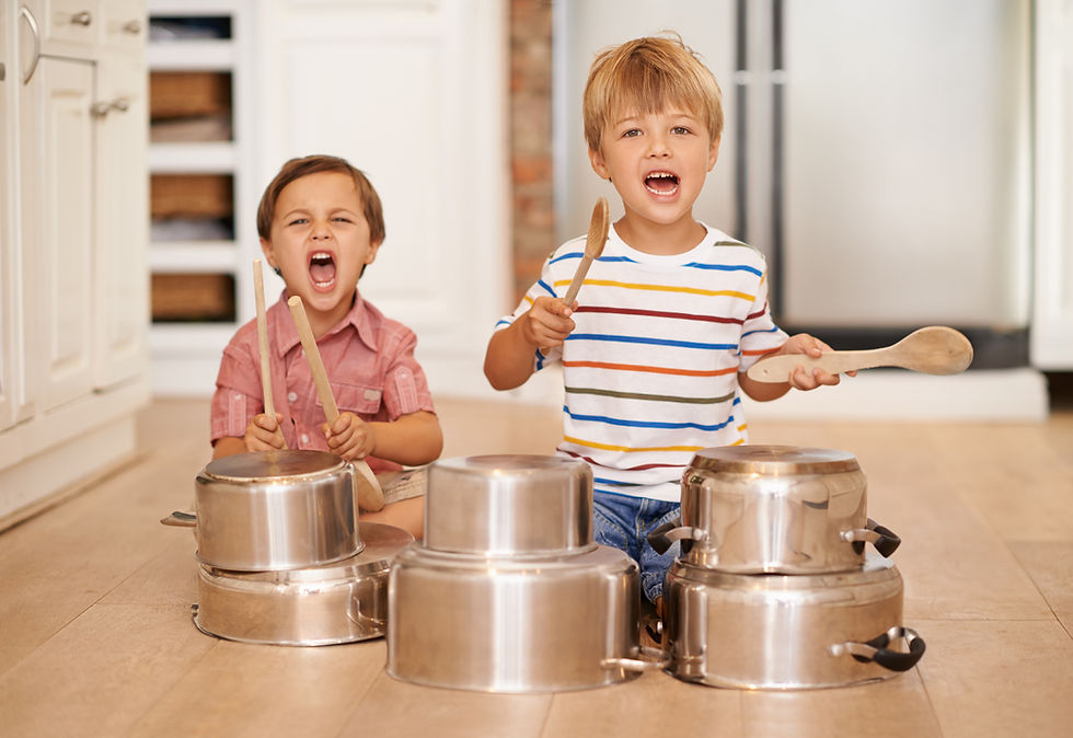 Two boys joyfully play drums with spoons on pots in a bright kitchen. One wears stripes, the other pink. Both appear energetic and playful.
