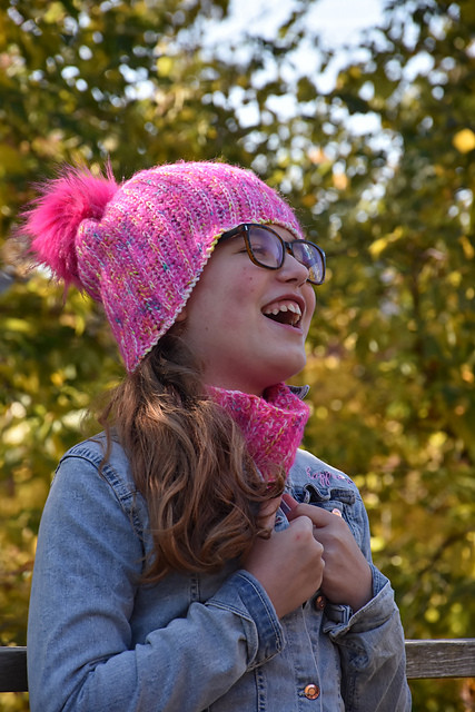 Une enfant avec des lunettes rit. Elle porte une tuque rose à pompon.