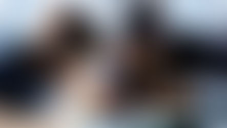 Two women working together in an automotive repair shop, wearing coveralls and using tools to fix a vehicle on a lift.