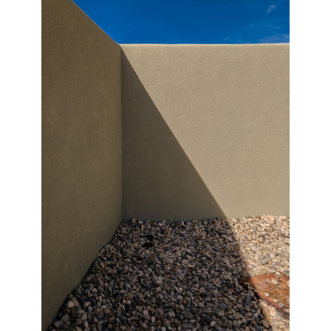 Minimalist outdoor corner wall with light stucco finish, desert rock landscaping, and strong diagonal shadows.