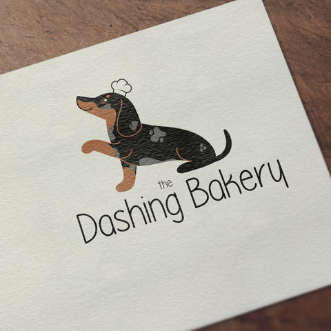 A digital mockup of a logo that features a dachshund with a chef hat on, posing with it's paw up. The words 'The Dashing Bakery' underneath show the name of the business that this logo was intended for. The logo is made to look like it is printed on a textured piece of paper that is laid on a wooden table.